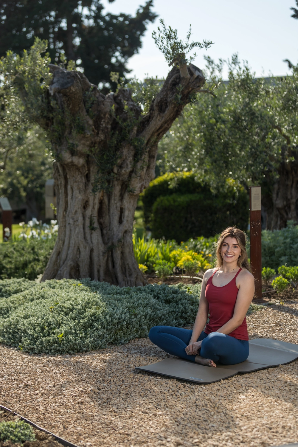 Yoga für Dich - Yoga Kurse - Eine lächelnde Frau in einem roten Tanktop und blauen Leggings sitzt im Schneidersitz auf einer Yogamatte in einem sonnigen Garten, hinter ihr ein großer, knorriger Olivenbaumstamm.