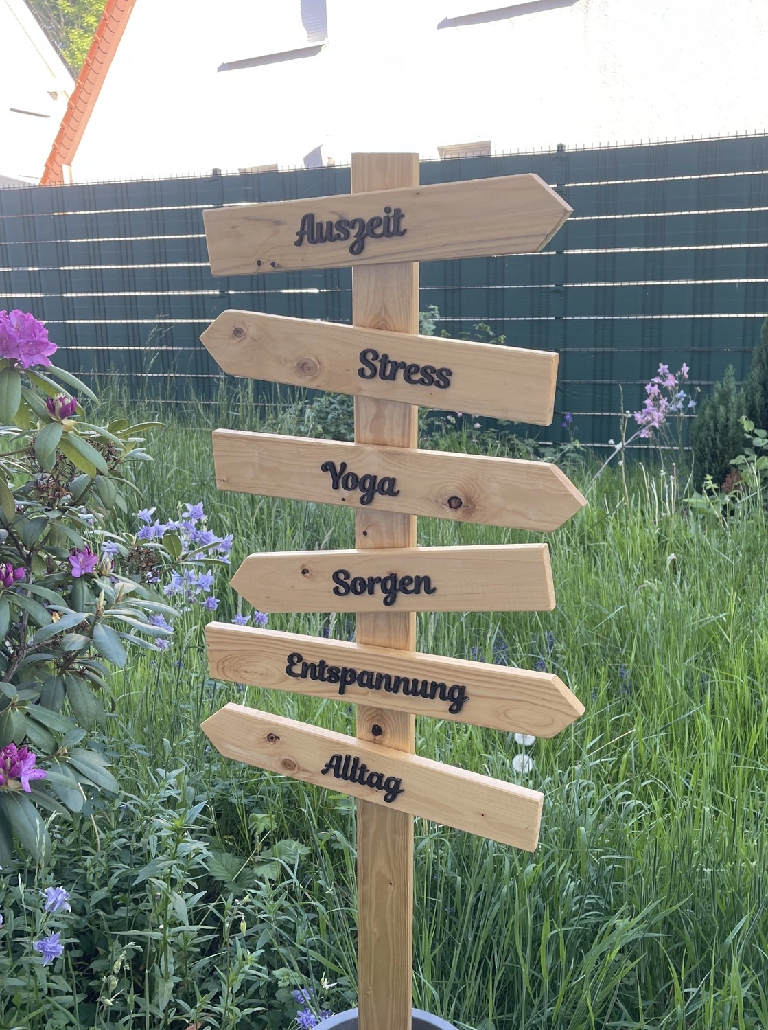 Yoga für Dich - Yoga Kurse - Ein hölzerner Wegweiser mit sechs Richtungspfeilen, die auf die Worte „Auszeit“, „Stress“, „Yoga“, „Sorgen“, „Entspannung“ und „Alltag“ in einer Gartenumgebung hinweisen.