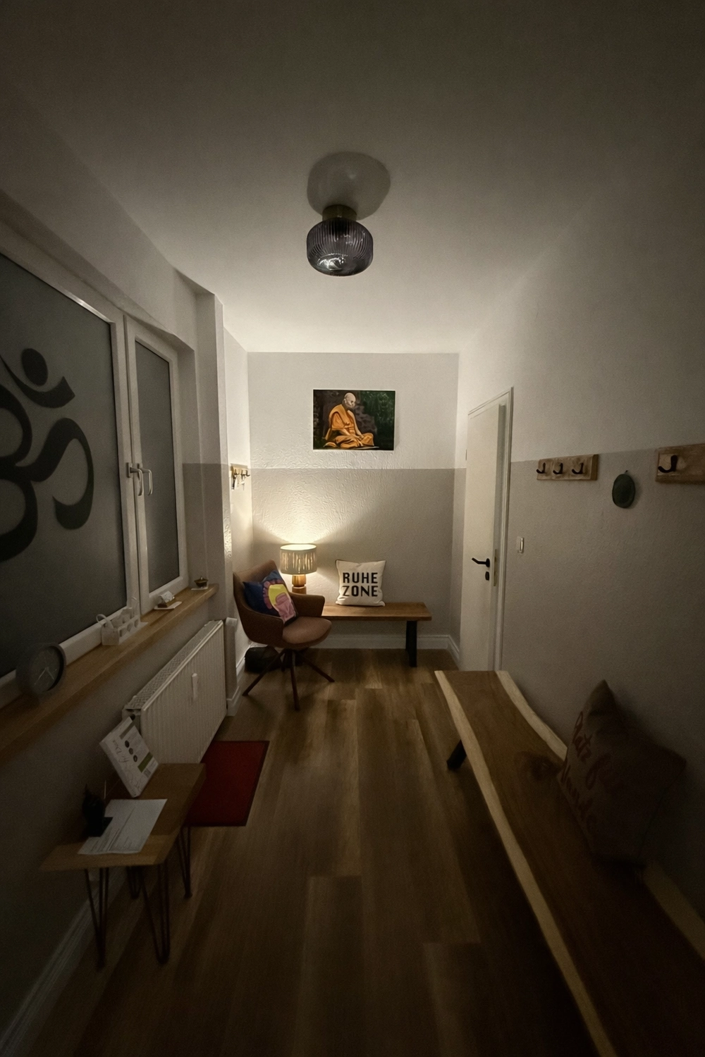 Yoga für Dich - Yoga Kurse - Ein gemütlicher, schwach beleuchteter Raum oder Flur, der als Meditations- oder Ruhebereich gestaltet ist und ein großes Om-Symbol an einem Fenster, ein Gemälde eines meditierenden Mönchs, Holzmöbel, darunter eine Bank mit einem Kissen mit der Aufschrift "RUHE ZONE" (Ruhezone), und hölzerne Kleiderhaken aufweist.