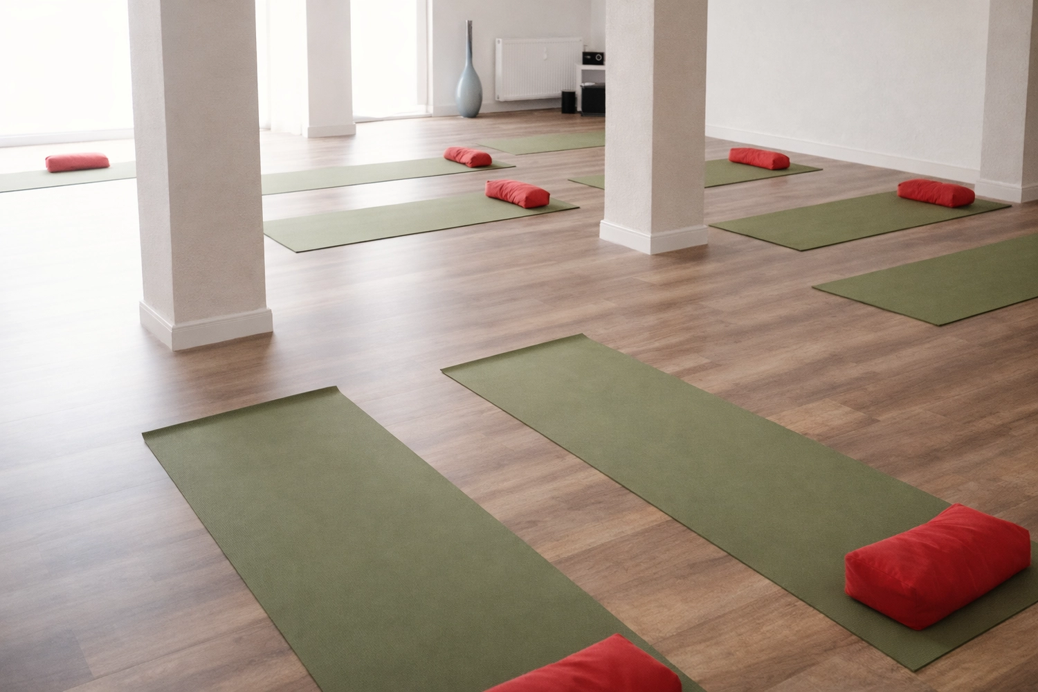 Yoga für Dich - Yoga Kurse - Ein helles, leeres Yogastudio mit Holzboden, in dem mehrere grüne Yogamatten und rote, rechteckige Kissen in Reihen angeordnet sind. Der Raum hat weiße Wände und weiße Säulen.