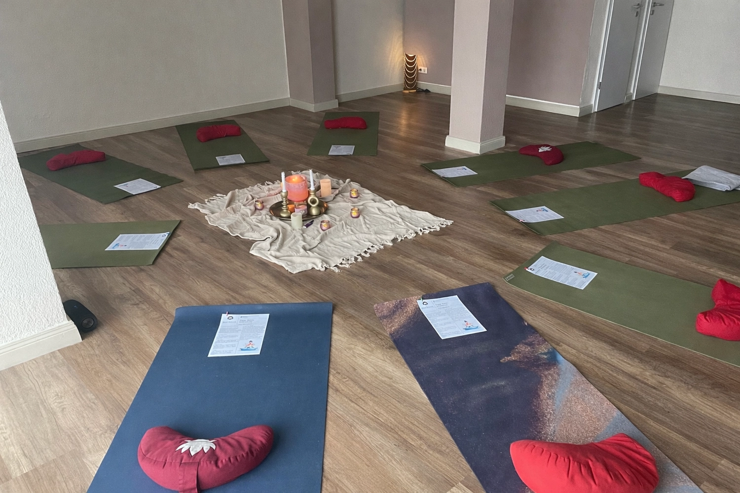 Yoga für Dich - Yoga Kurse - Ein helles, leeres Yogastudio mit Holzboden, in dem mehrere grüne Yogamatten und rote, rechteckige Kissen in Reihen angeordnet sind. Der Raum hat weiße Wände und weiße Säulen.