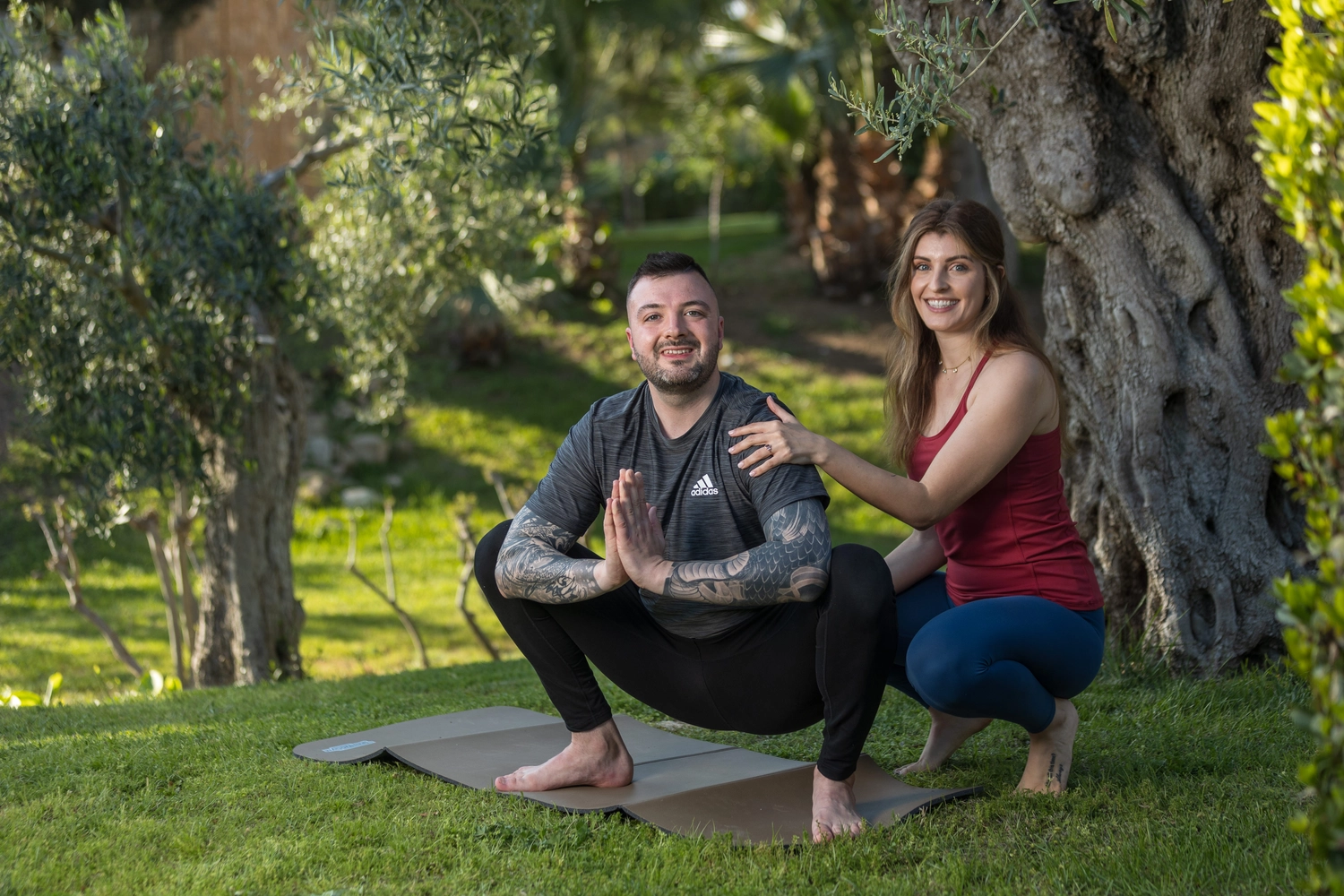 Yoga für Dich - Yoga Kurse - Ein Mann und eine Frau sitzen im Freien inmitten der Natur und lächeln in die Kamera. Der Mann hockt mit gefalteten Händen, die Frau sitzt neben ihm und hat ihre Hand auf seine Schulter gelegt.